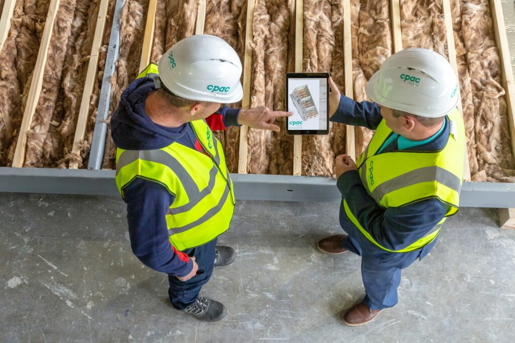 Two construction workers in safety vests and hard hats look at a tablet together while standing indoors near exposed insulation and wooden beams on the floor.