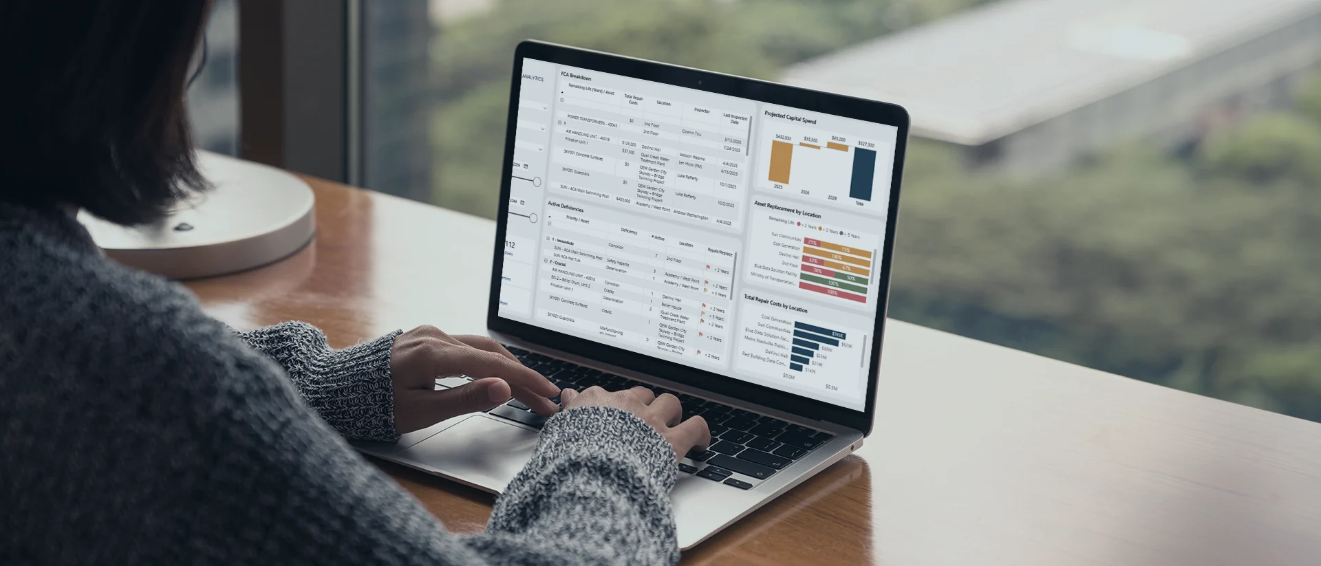 A person in a gray sweater uses a laptop on a wooden table by a window. The screen displays charts, graphs, and tables with financial or business data.