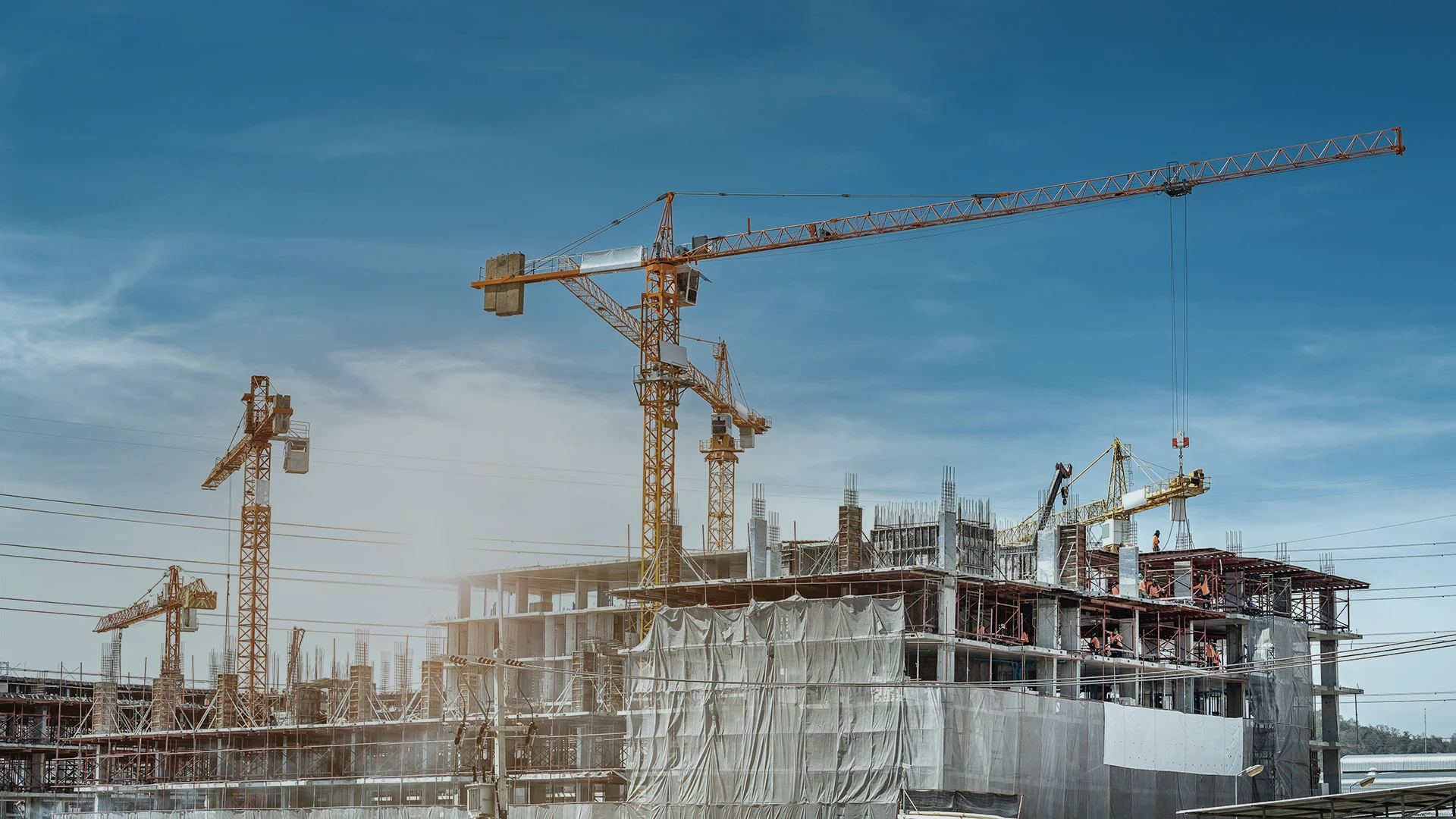 Construction site with multiple cranes and a partially built multi-story building covered in scaffolding and netting, set against a clear blue sky.
