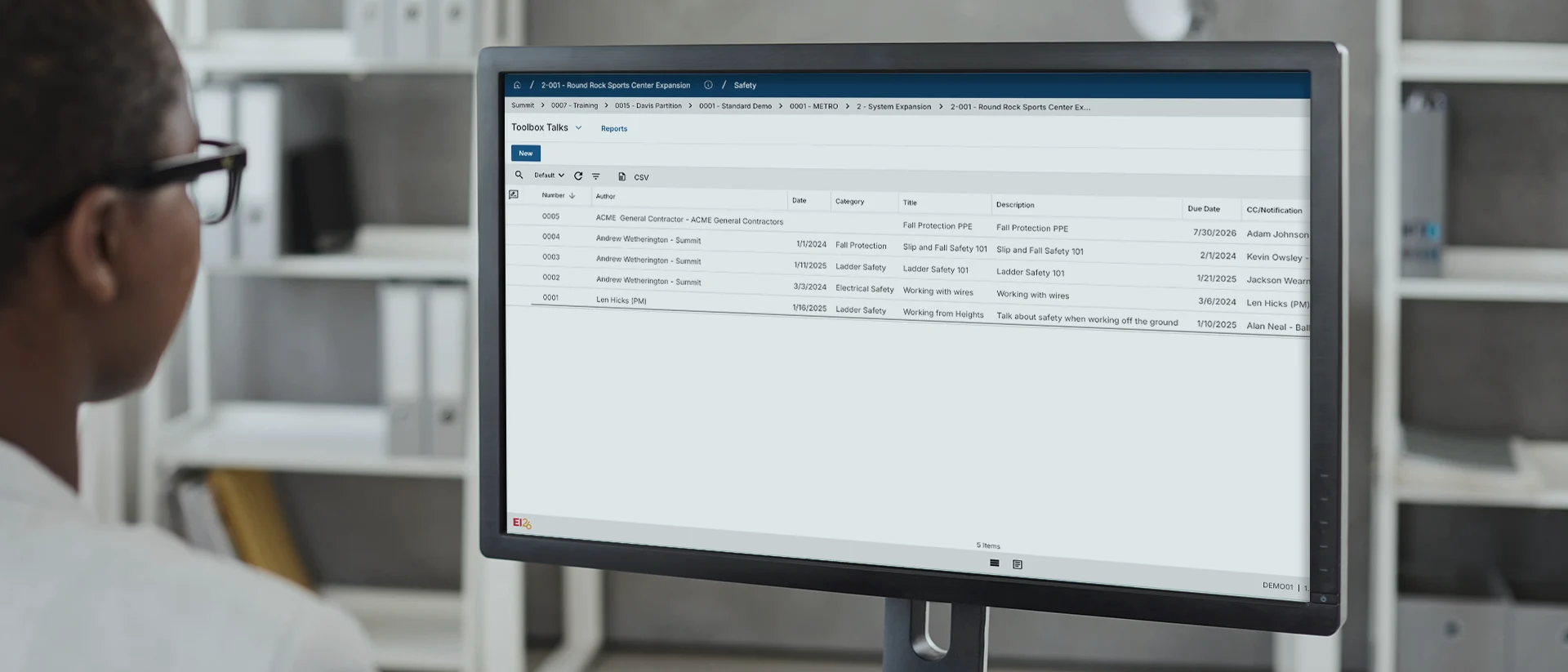 A computer monitor displays a task management dashboard with several listed tasks, their statuses, categories, deadlines, and completion dates in an office setting. Shelves with binders are visible in the blurred background.