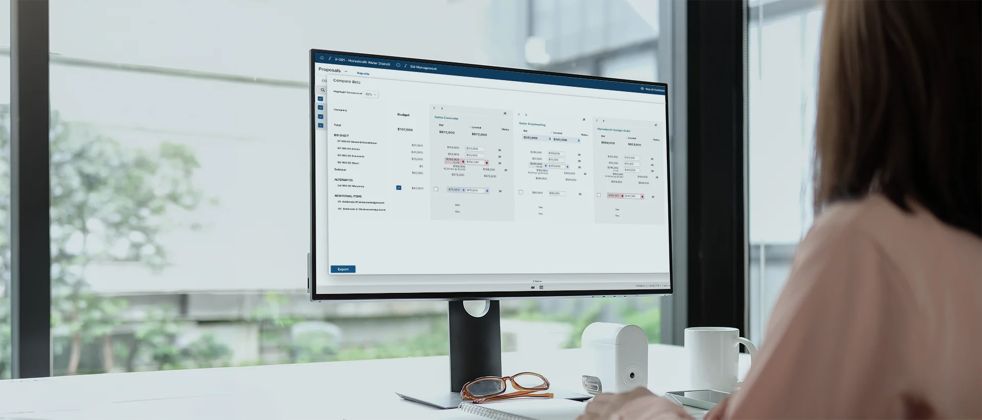 A person sits at a desk using a computer with a large monitor displaying data tables and filters in a web application, in a bright office with large windows.