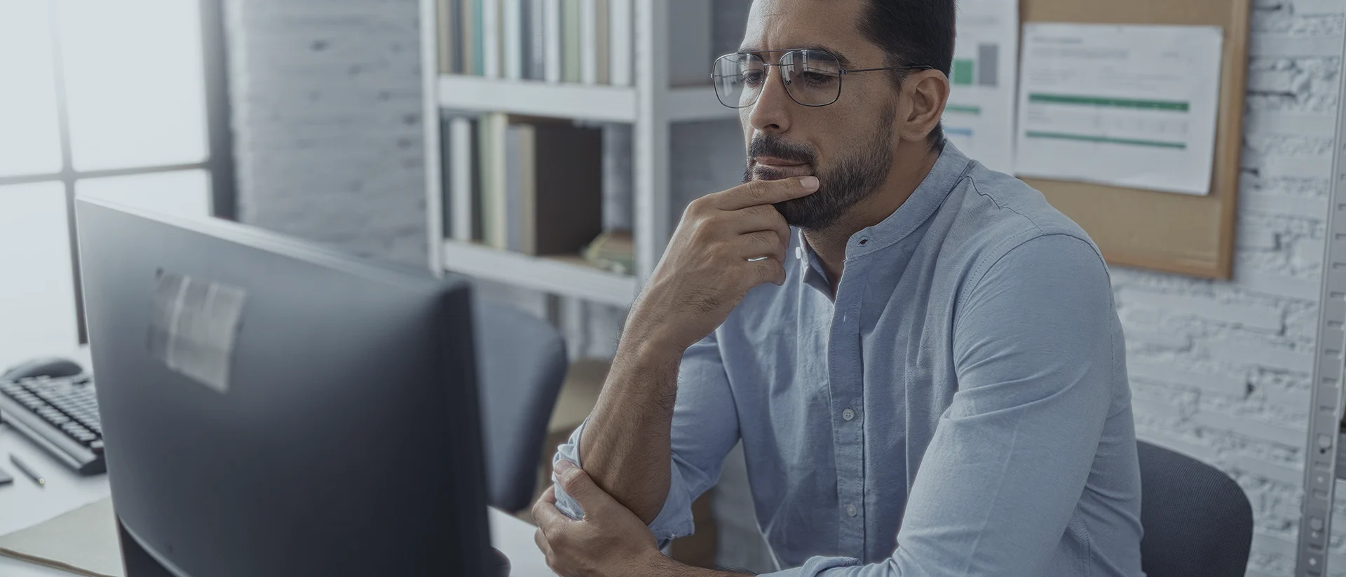 Man wearing glasses and a light blue shirt sits at a desk, looking thoughtfully at a computer monitor in a modern office with shelves and documents in the background.