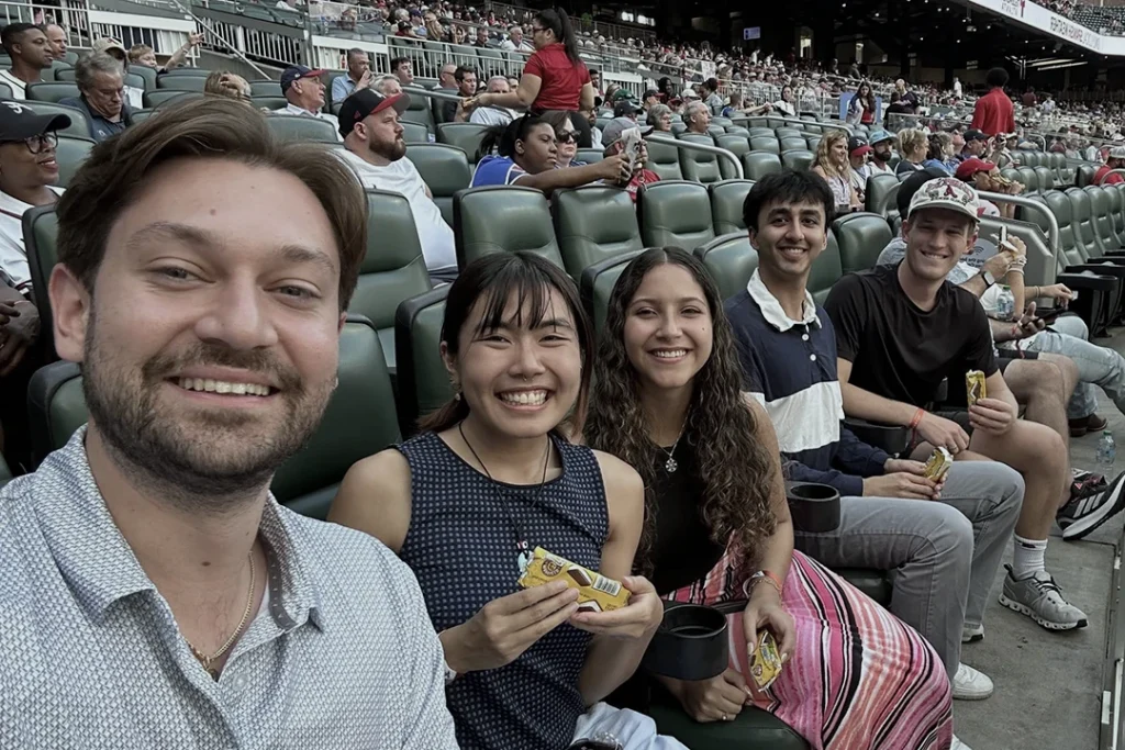 Five people sit smiling in stadium seats, holding snacks. The stands behind them are filled with other spectators, suggesting they are at a sporting event.