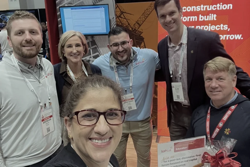 Six people pose and smile at a business expo booth, wearing badges and business or casual attire. A woman in front holds a certificate. A red display with text about construction is visible in the background.