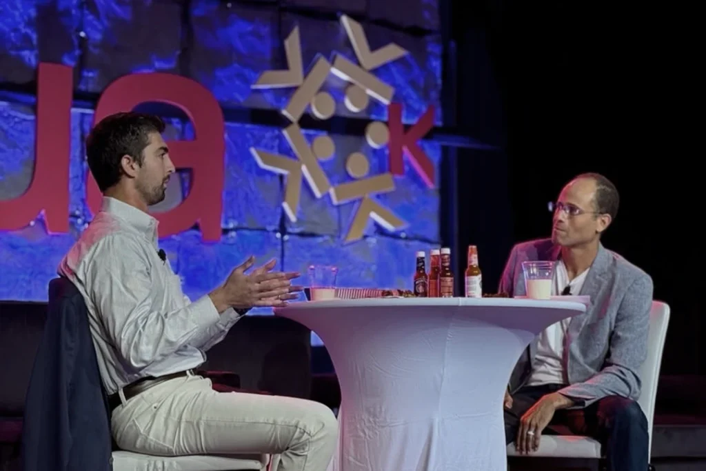 Two men sit at a round table on a stage, engaged in conversation. Bottles and a water pitcher are on the table. A backdrop with red and yellow symbols is behind them.