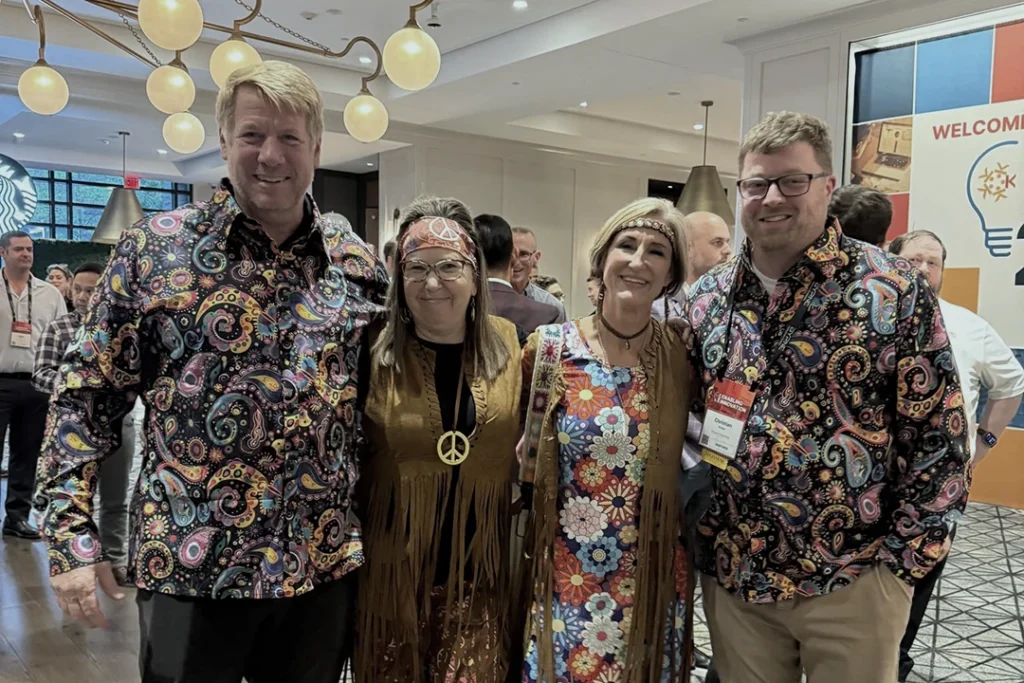 Four adults dressed in colorful, 1960s-inspired outfits pose and smile together at an indoor social event, surrounded by other people in the background.