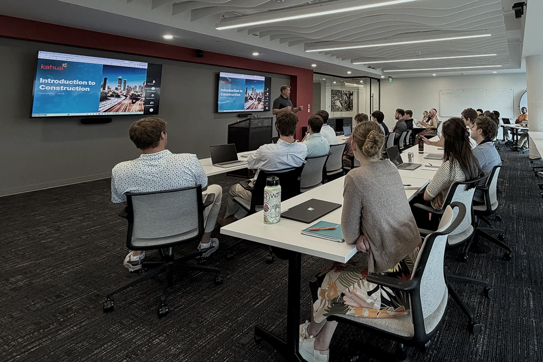 A group of people sit at desks in a modern classroom, watching a presenter at the front. A screen displays a slide titled Introduction to Construction. Laptops and notebooks are on the desks.