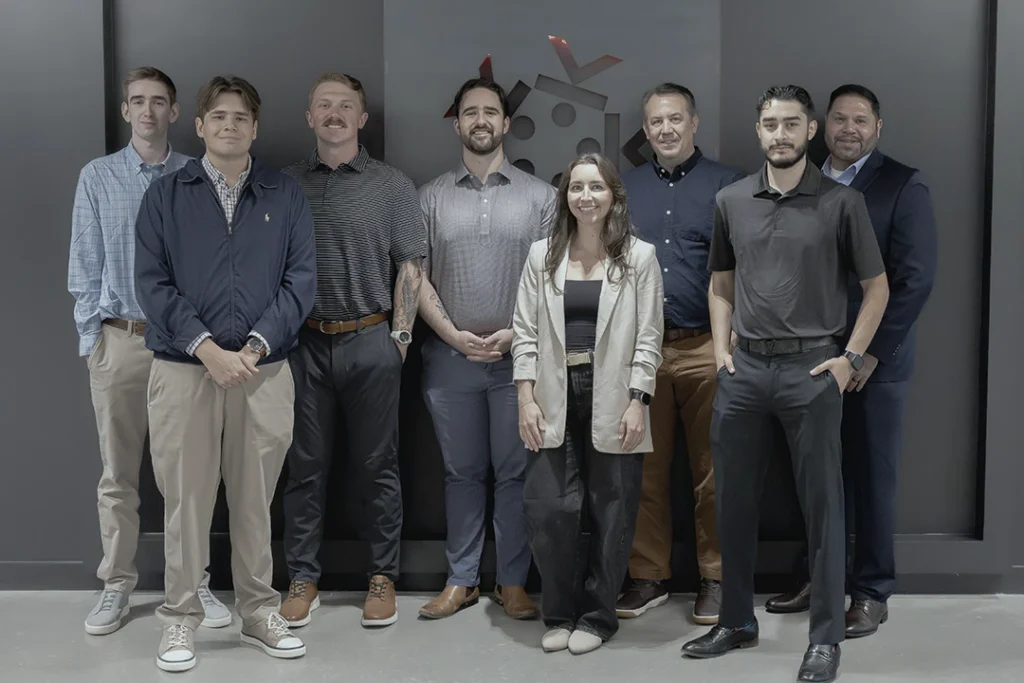Eight people, seven men and one woman, stand in front of a gray wall with a logo. They are dressed in business casual attire and are smiling at the camera.