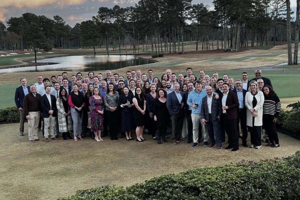 A large group of people pose together outdoors on a golf course at sunset, with trees, a pond, and green grass in the background. Most are dressed in business or semi-formal attire and are smiling at the camera.