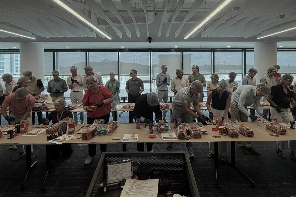 A group of people wearing hair nets assemble sandwiches at long tables in a modern, bright room with large windows. Various sandwich ingredients and bread are visible on the tables.