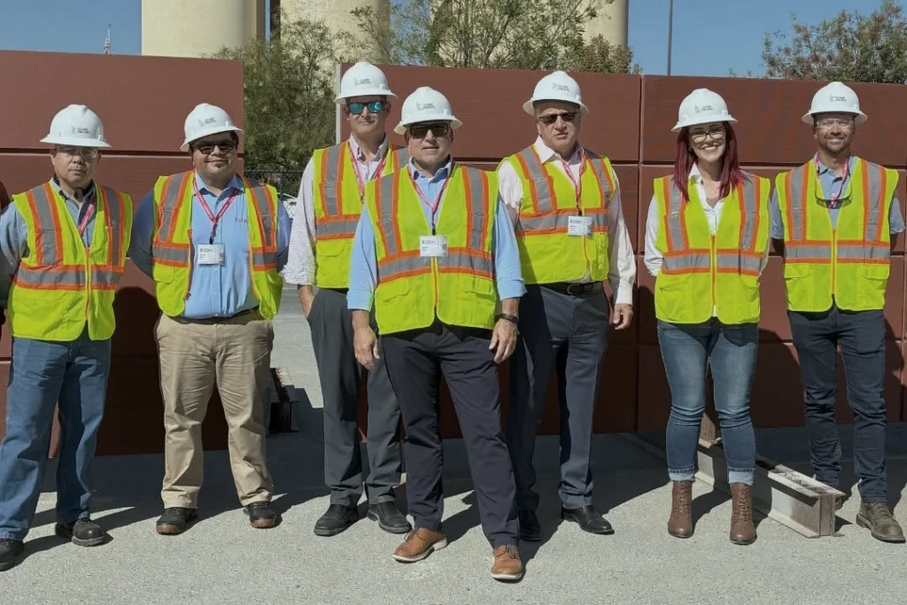 Seven people wearing hard hats, safety vests, and ID badges stand outdoors in front of a brown structure, posing for a group photo on a sunny day.