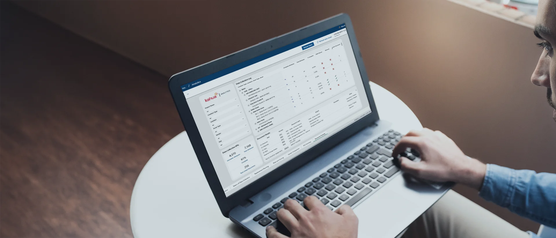 A person uses a laptop on a white table, viewing a dashboard with charts, graphs, and navigation menus on the screen. The environment is well-lit with natural light from a nearby window.