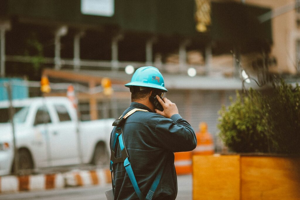 A construction worker wearing a blue hard hat and safety harness talks on a cell phone at a job site.