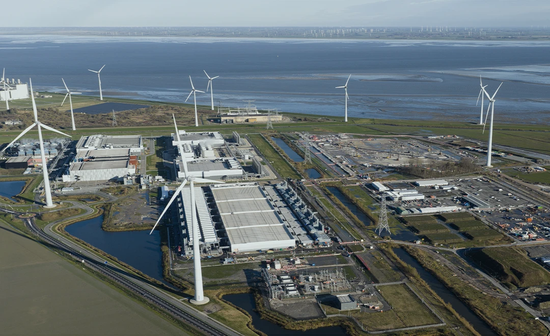 Aerial view of an industrial area near a coastline with several large wind turbines, factory buildings, parking areas, roads, and water channels visible.