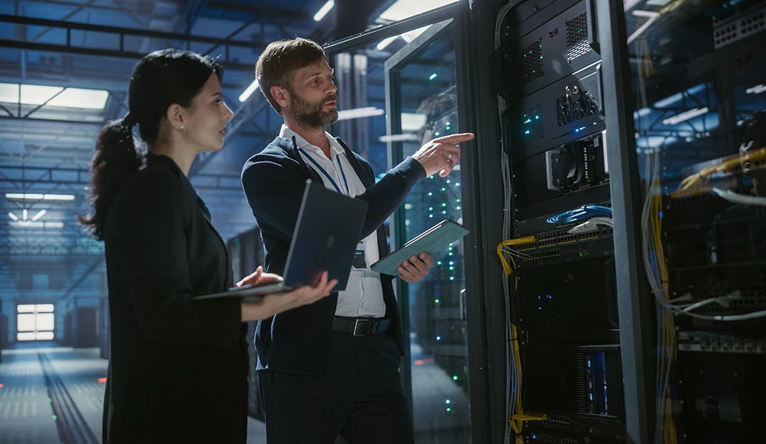 Two IT professionals stand in a server room, inspecting servers. One holds a laptop, the other holds a tablet and points toward the server rack. Blue lights and cables are visible in the high-tech data center environment.