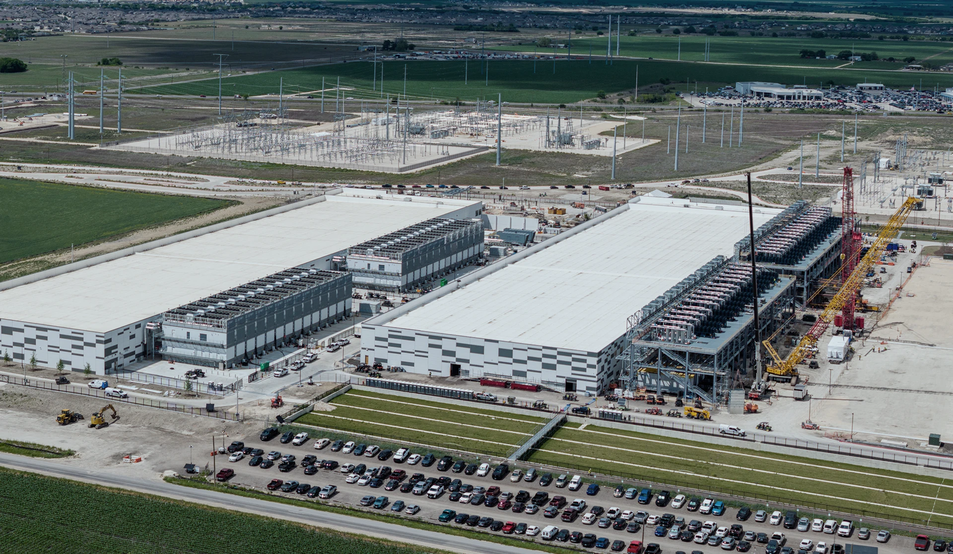Aerial view of a large industrial facility with two main buildings, cooling units, cranes, and a parking lot filled with cars, surrounded by green fields and power lines.