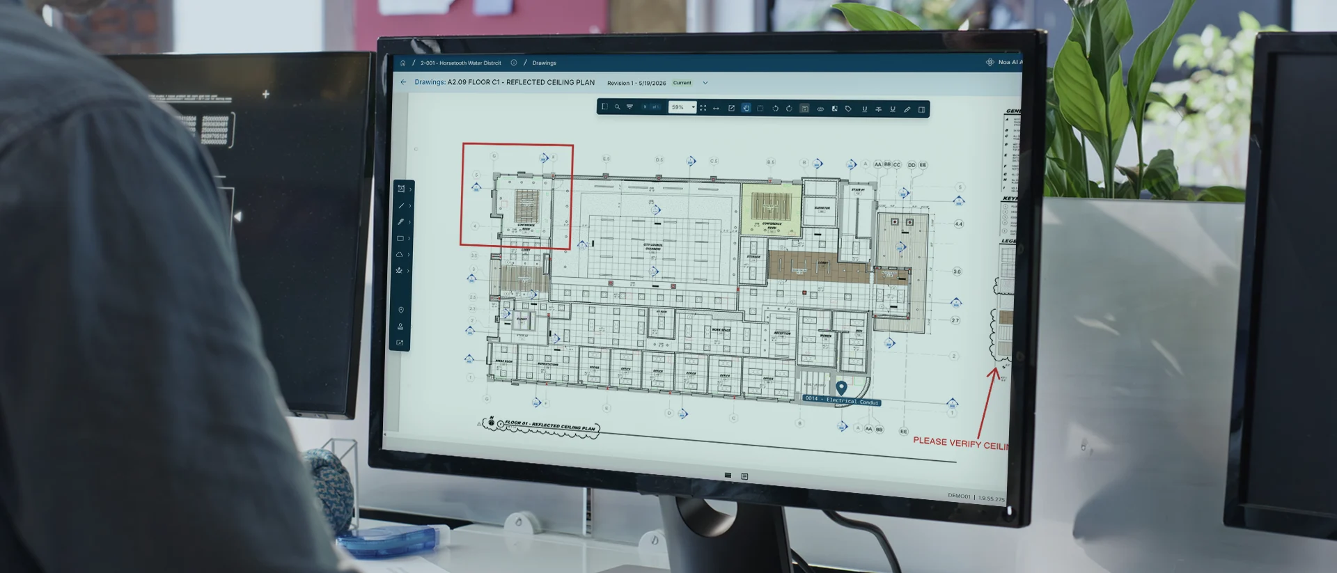 A computer monitor displays a detailed architectural floor plan with various annotations and symbols. The plan has a red highlighted section and a note that says “PLEASE VERIFY LOCATION.” Plants and office items are nearby.