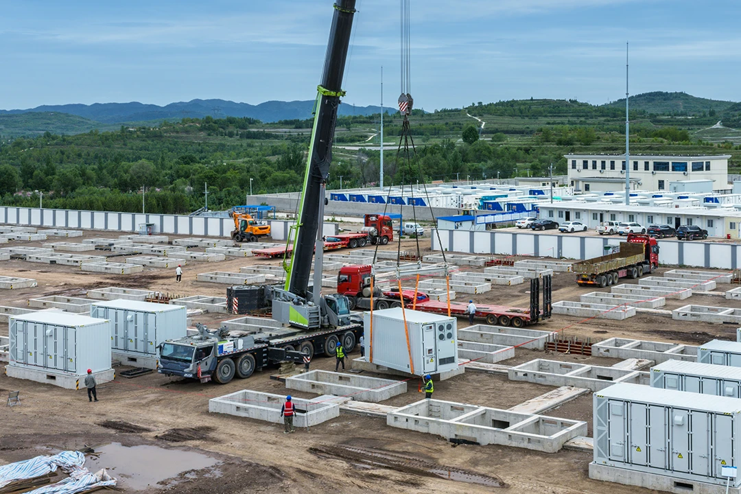 Construction site with cranes placing modular units onto concrete foundations, trucks and workers scattered across the area, surrounded by prefab buildings and distant green hills under a cloudy sky.