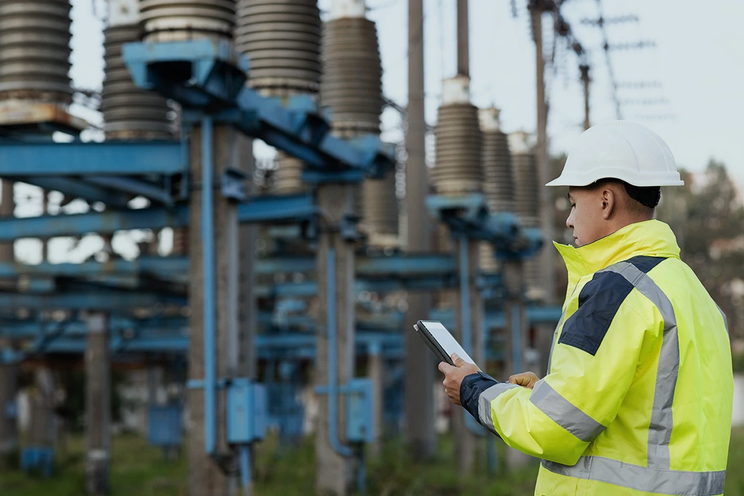 A worker in a high-visibility jacket and hard hat inspects electrical equipment outdoors, holding a tablet and standing among blue metal structures at a power facility.