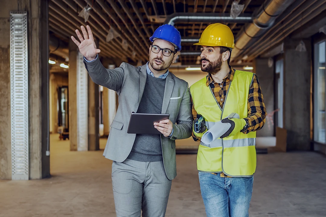 Two men wearing hard hats stand in an unfinished building; one in a suit gestures while holding a tablet, the other in a safety vest holds blueprints and looks where the first man is pointing.