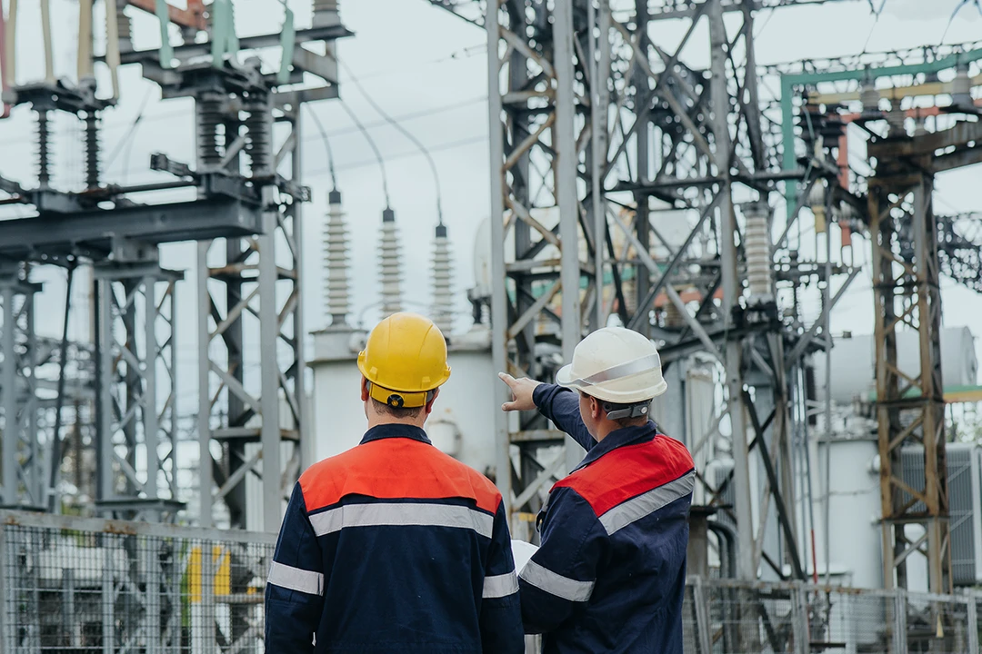 Two workers in safety gear and hard hats stand at an electrical substation. One worker points toward equipment while the other observes high-voltage structures and transformers.
