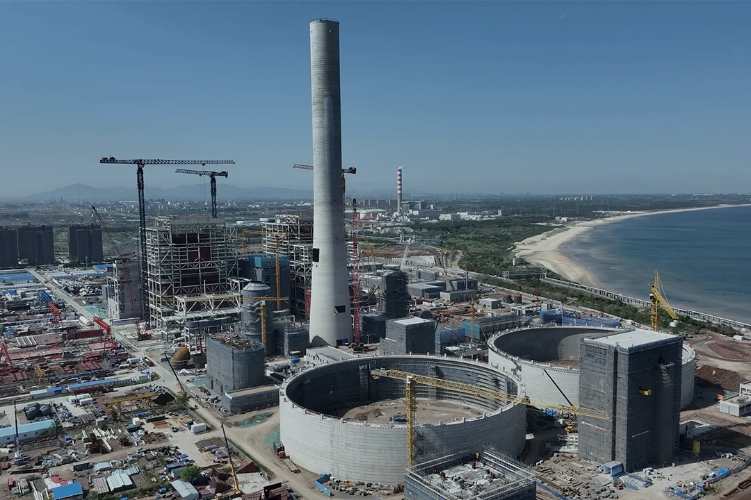 Aerial view of a large coastal construction site with cranes, cylindrical structures, a tall smokestack, and nearby buildings. The sea and sandy coastline are visible on the right.