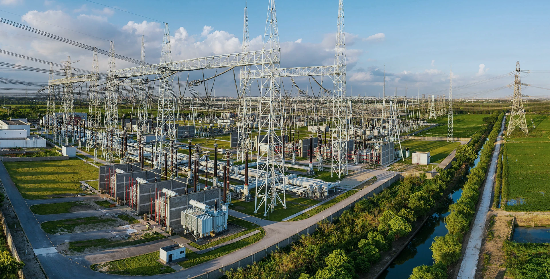 A large electrical substation with numerous transmission towers, power lines, equipment, and green fields, set under a partly cloudy sky.