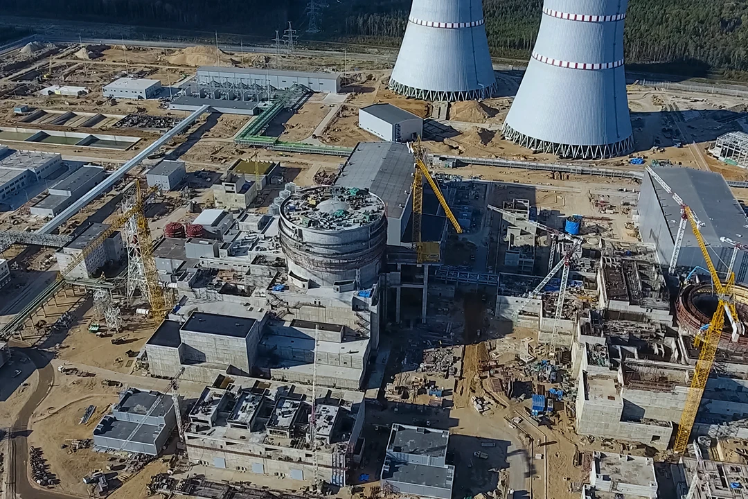 Aerial view of a large construction site with multiple cranes, buildings, and two tall cooling towers in the background, possibly part of a power plant.