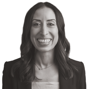Smiling woman with long dark hair, wearing a blazer over a light top, facing the camera against a plain white background.