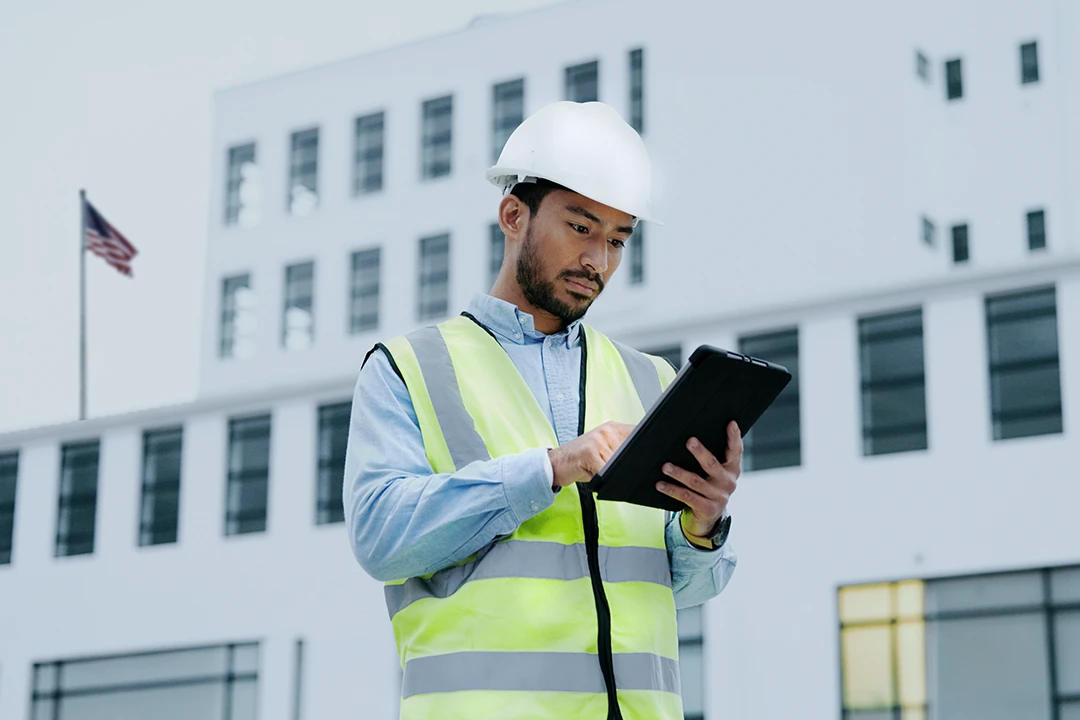 A construction worker in a hard hat and safety vest uses a tablet outside a modern building with an American flag in the background.
