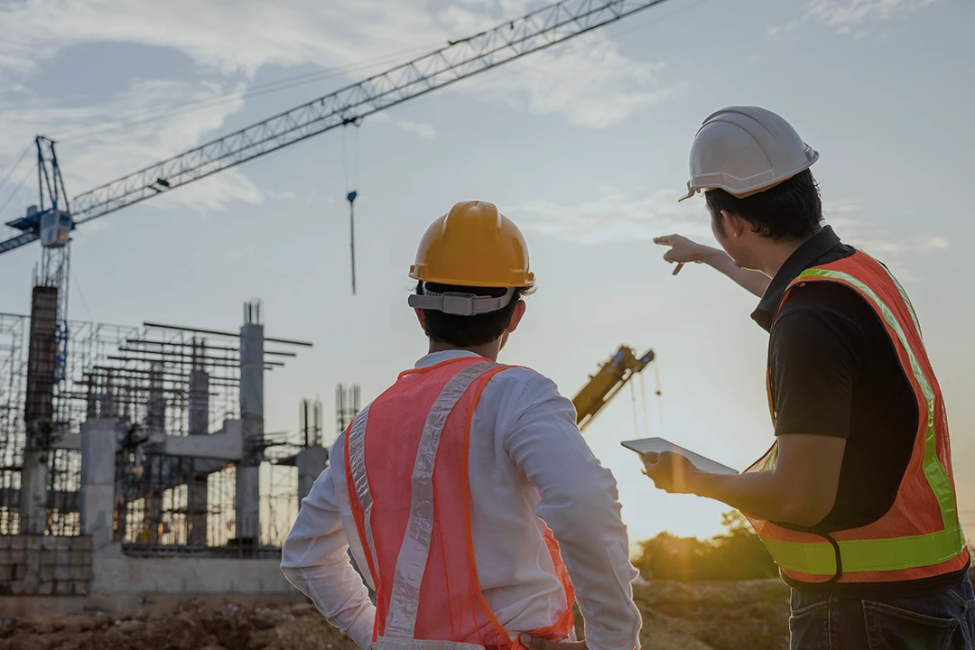 Two construction workers in safety vests and helmets discuss a building project at a construction site, with cranes and structural steel visible in the background during sunset.