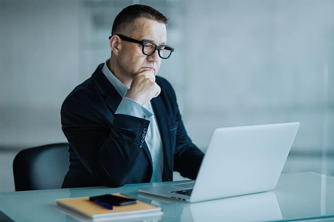 A man in a suit and glasses sits at a desk, looking thoughtfully at a laptop. A notebook and a phone are on the table in front of him. The background is blurred and neutral.