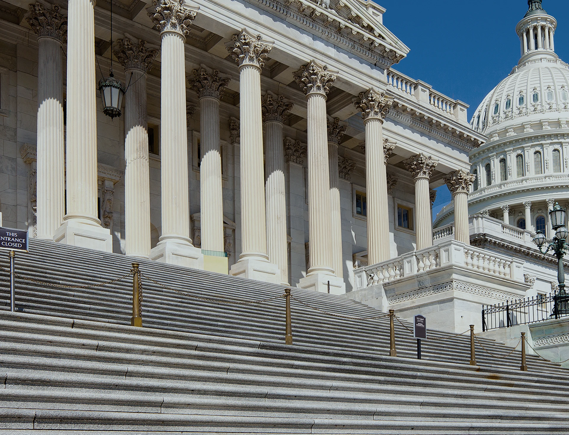 Wide staircase leading up to the columns of a large white government building, with part of a dome visible in the background under a clear blue sky.