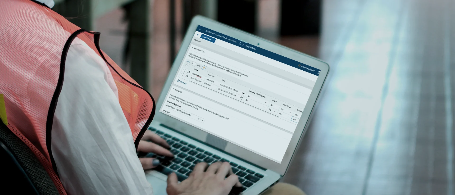 A person wearing a safety vest types on a laptop displaying a software interface with tables and form fields.