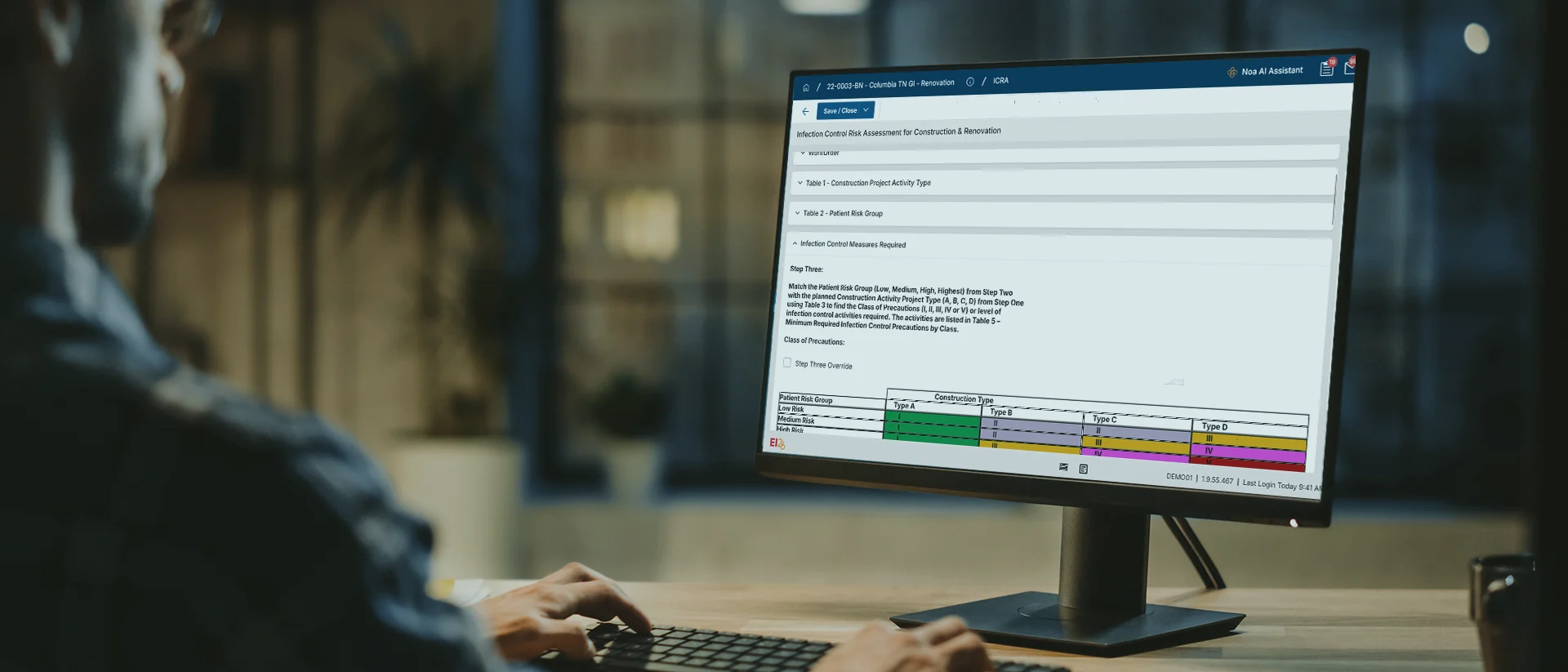 A person sits at a desk, using a desktop computer with a large monitor displaying a detailed chart and text in a web application interface.
