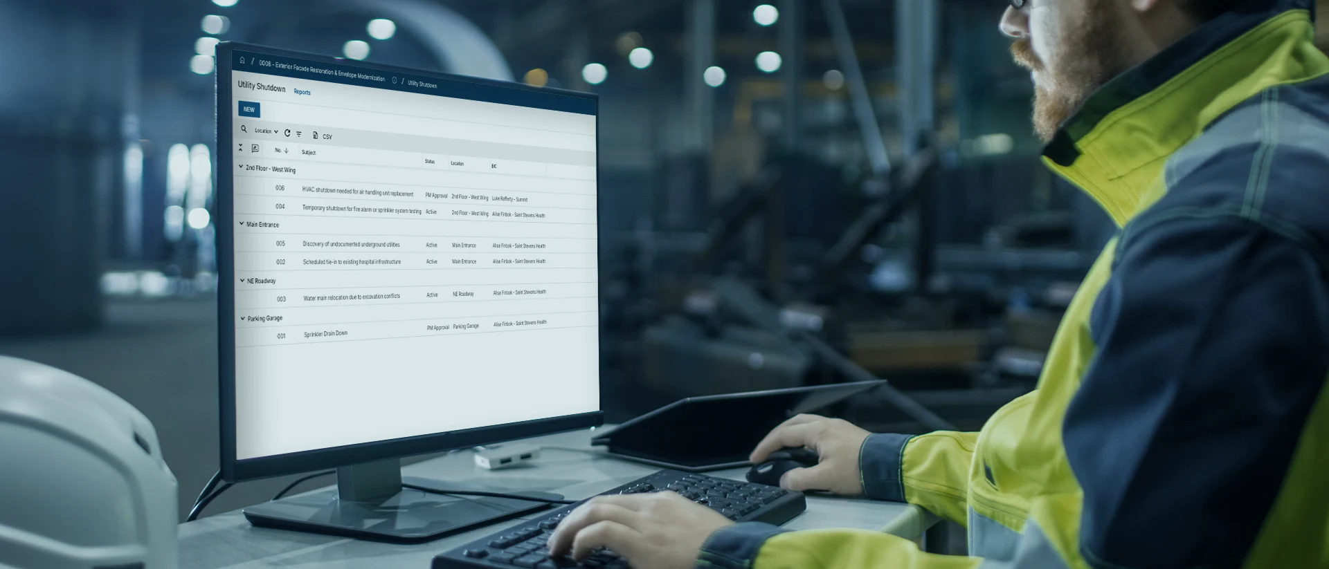 A person in a high-visibility jacket works at a computer, viewing a dashboard in an industrial setting. The background is blurred, showing machinery and blue lights.