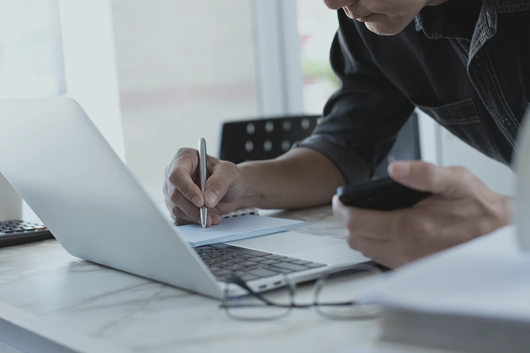 A person writes on a notepad while holding a smartphone, with an open laptop and eyeglasses nearby on a desk in a bright room.