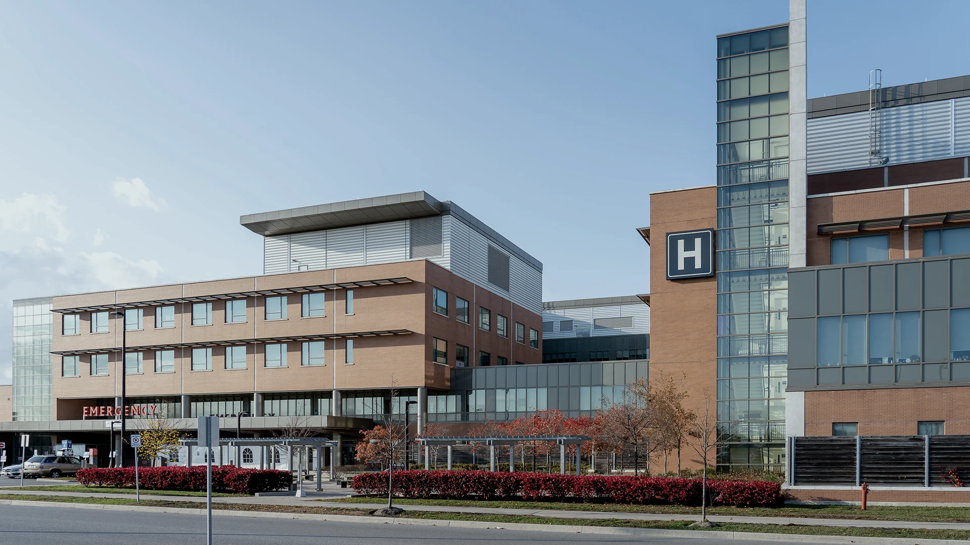 Modern hospital building with large glass windows and a brick exterior. An emergency entrance is visible on the left, and a prominent H sign denotes the hospital. Small trees with red leaves line the front.