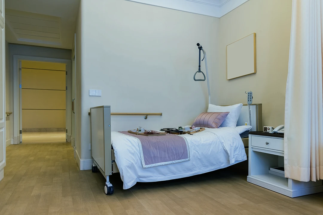 A hospital room with a single bed, light pink bedding, a tray with food, a bedside table, and medical equipment, all set against beige walls and wooden flooring.