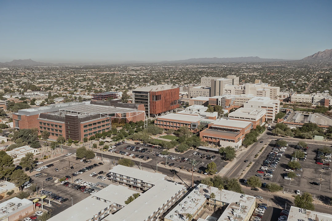Aerial view of a large urban campus with multiple modern buildings, parking lots, and scattered trees, set against a backdrop of cityscape and distant mountains under a clear blue sky.