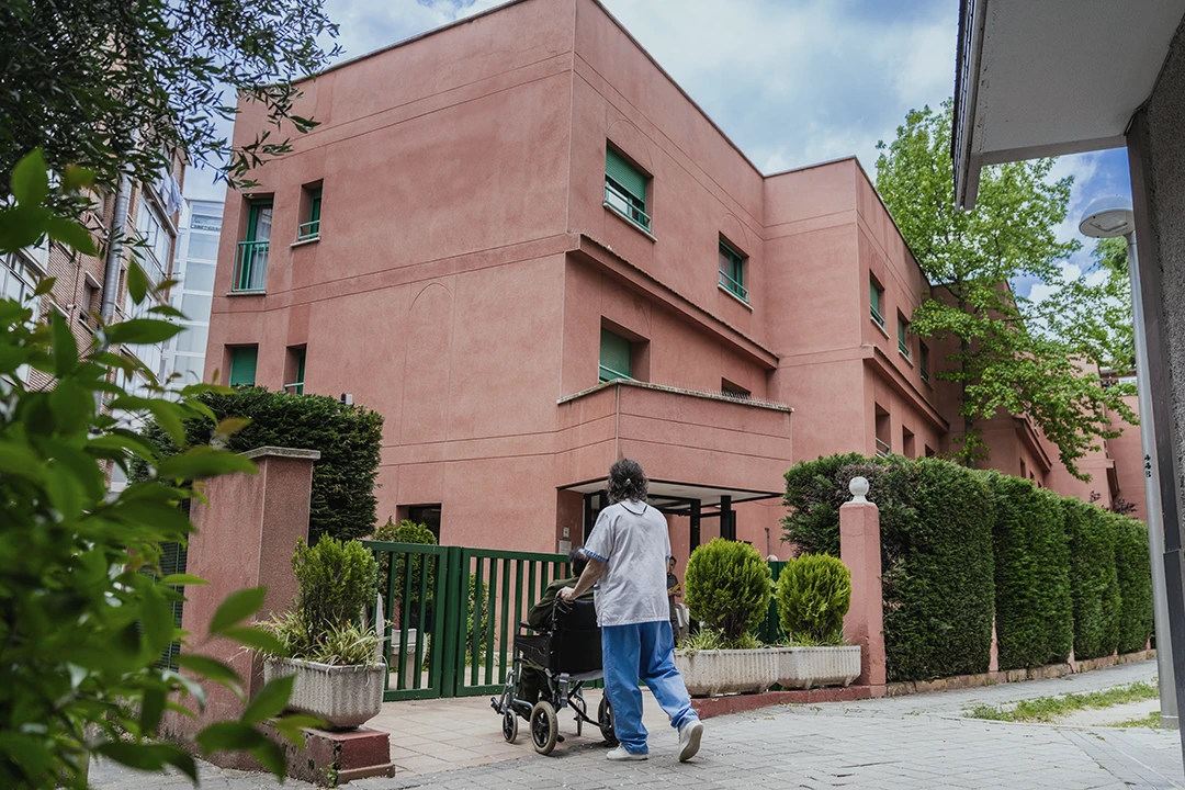 A person pushes a wheelchair toward the entrance of a pink, two-story building with green window shades and surrounding greenery on a partly cloudy day.