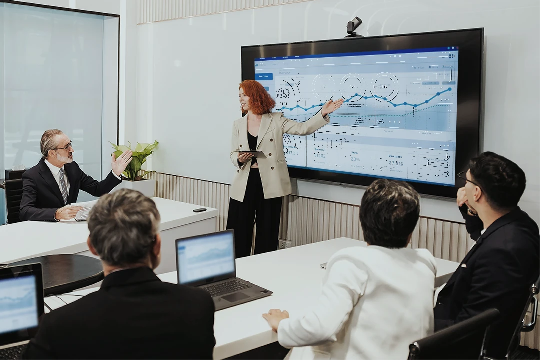 A woman stands and presents data on a large screen to four seated colleagues in a modern conference room. Laptops are on the table, and everyone is focused on the presentation.