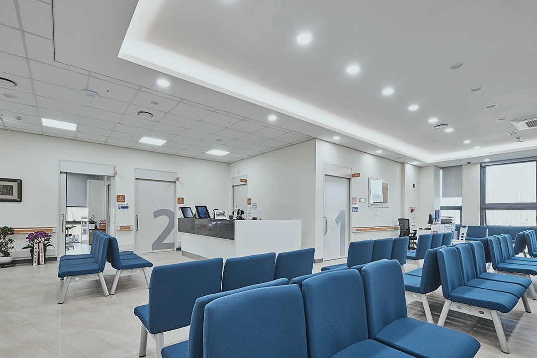 Modern medical waiting room with blue chairs, a white reception desk, and numbered doors. Bright lighting and large windows create a clean, welcoming atmosphere.