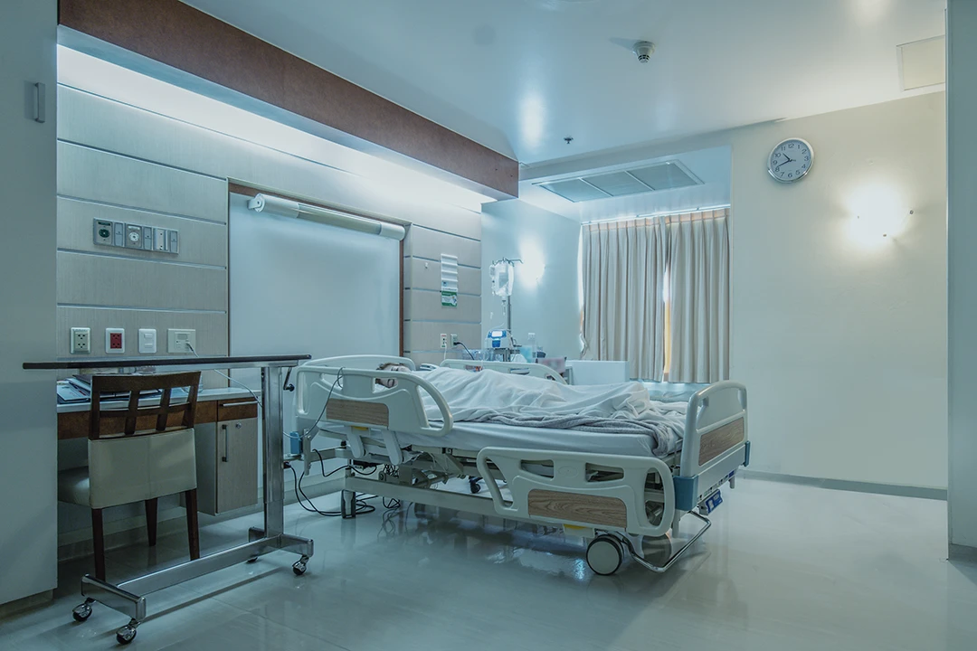A hospital room with a patient lying in a bed, covered by a blanket. Medical equipment, a chair, and a table are nearby. Light enters through curtained windows; a clock is on the wall.