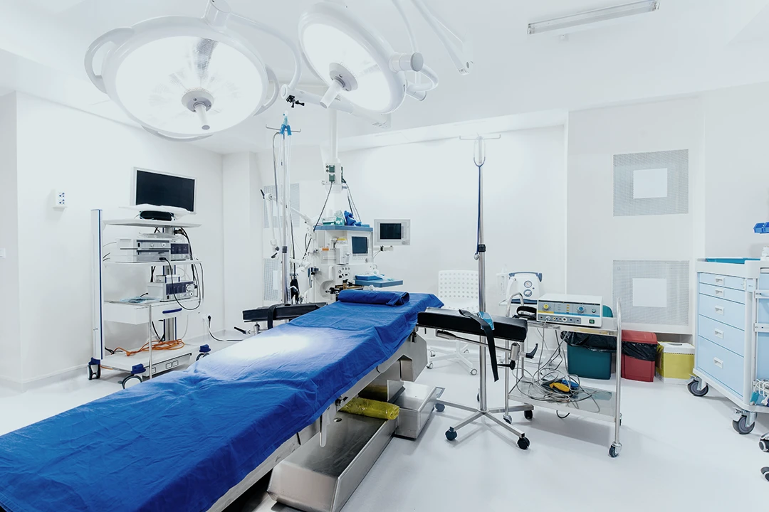 A clean, modern operating room with a blue-covered surgical table, medical equipment, monitors, bright overhead lights, and storage cabinets in a white, sterile environment.