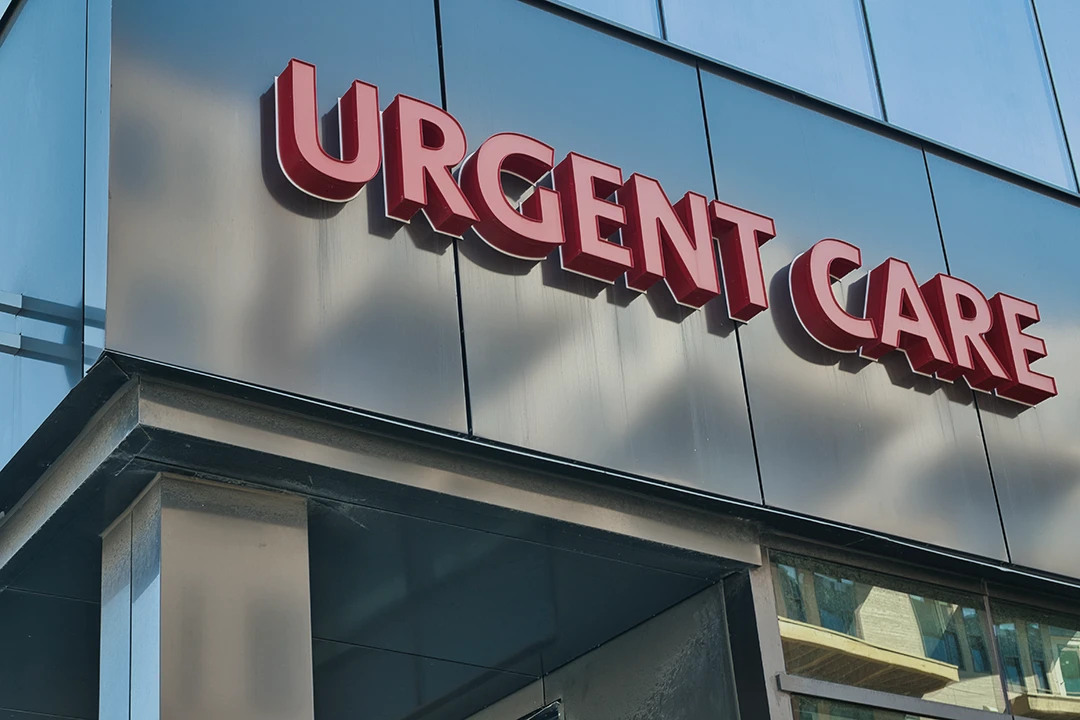 Red URGENT CARE sign on the exterior of a modern glass building, reflecting nearby structures in the windows below the sign.