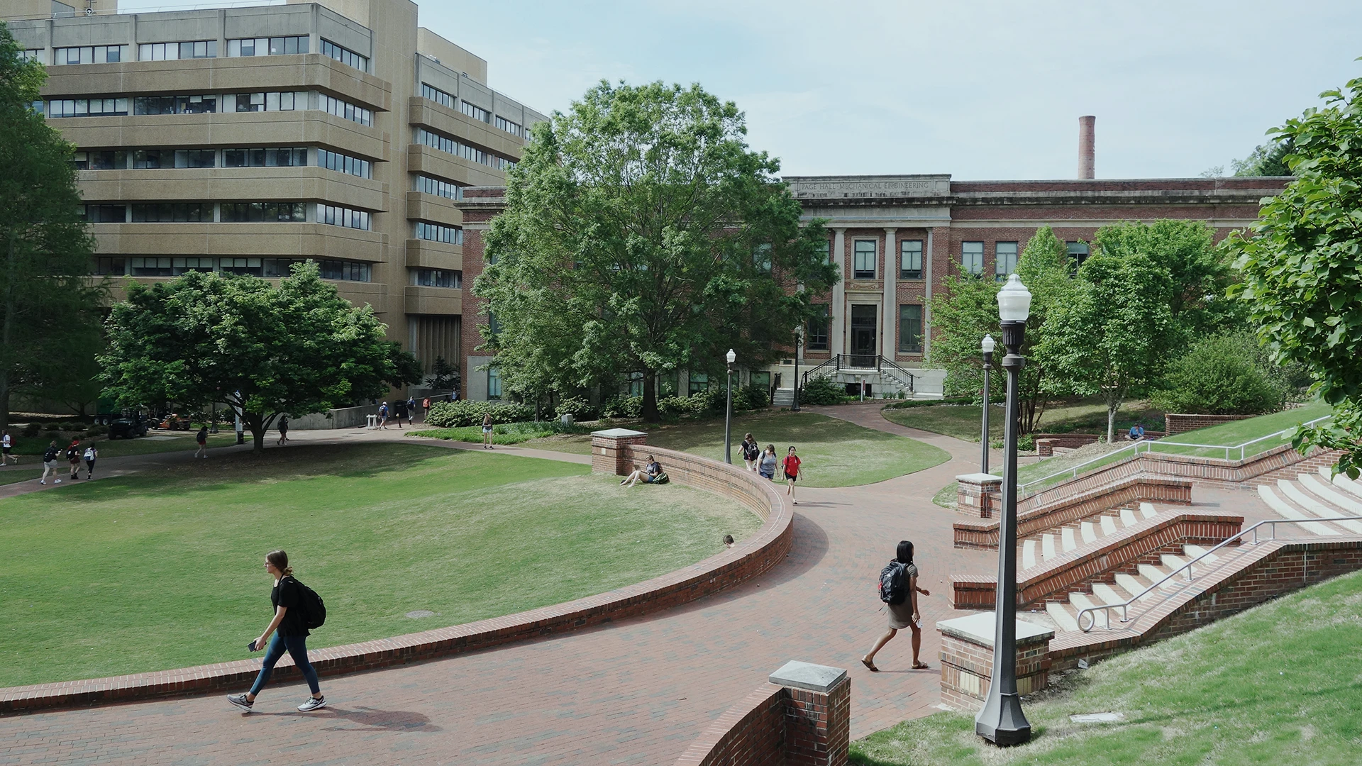 A college campus scene with students walking on curved brick paths, surrounded by green lawns and trees, near multi-story academic buildings.