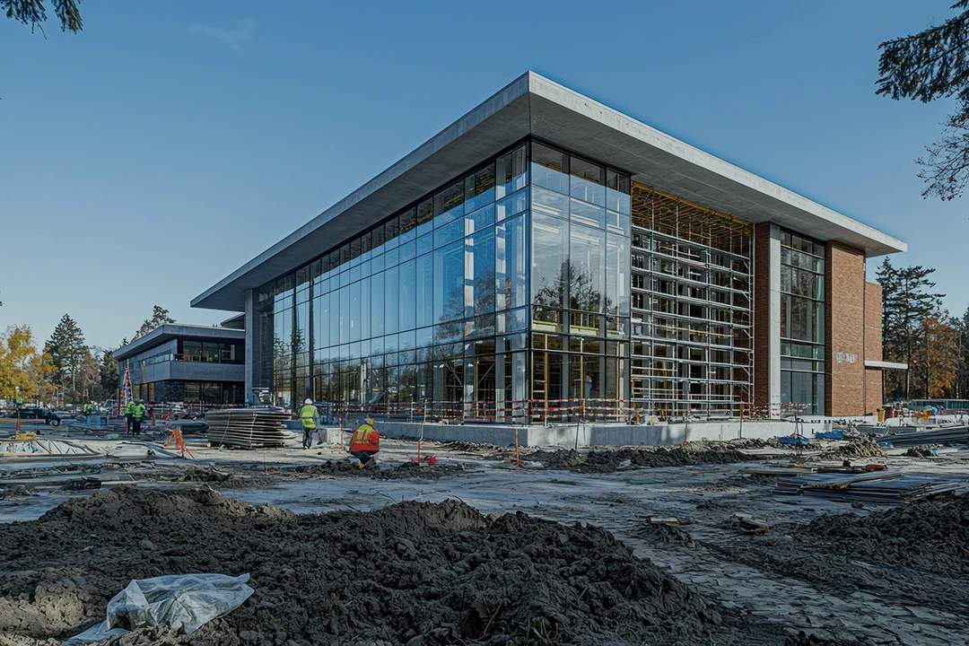 A modern building with large glass windows under construction, surrounded by dirt, workers in safety vests, and construction materials. Another similar building is visible in the background, with trees and a blue sky.