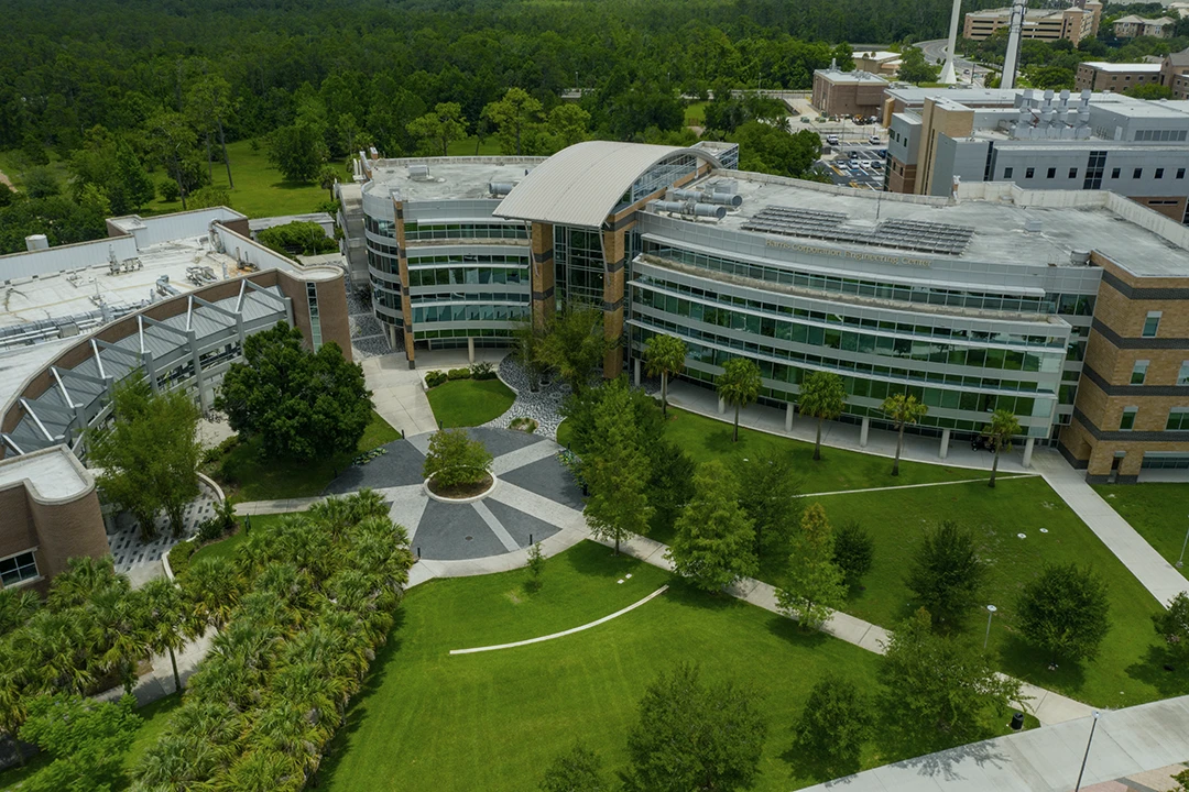 Aerial view of a modern, multi-story campus building with curved glass walls, surrounded by green lawns, trees, and walkways, set against a backdrop of wooded areas.