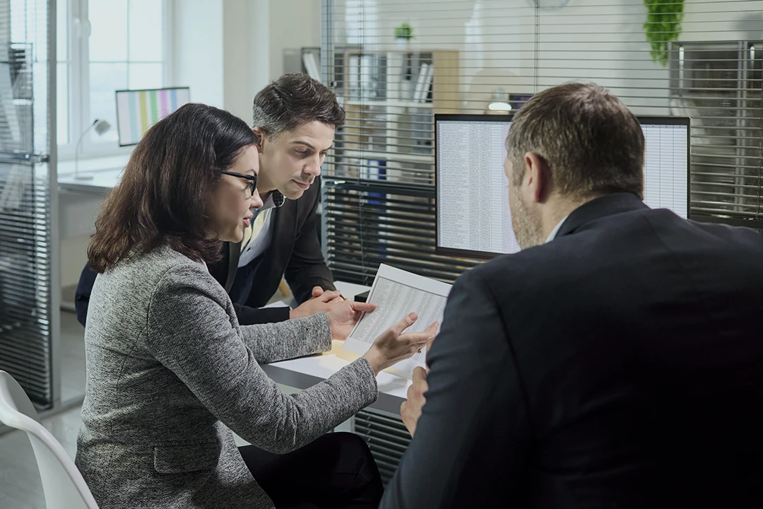 Three people in business attire sit at a desk, examining documents together in a modern office with computers and blinds in the background.